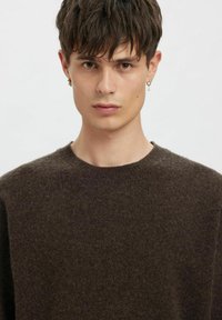 Rjavo pulover z mehko teksturo in okroglim izrezom. Modelka ima kratke lase in zlato obročno uhan, z nevtralnim izrazom na obrazu.