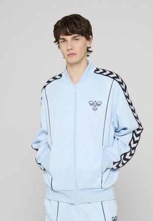 Lichtblauwe jas met ritssluiting gemaakt van zacht materiaal; heeft zwarte zigzagstrepen op de schouders en een klein grafisch logo op de borst.
