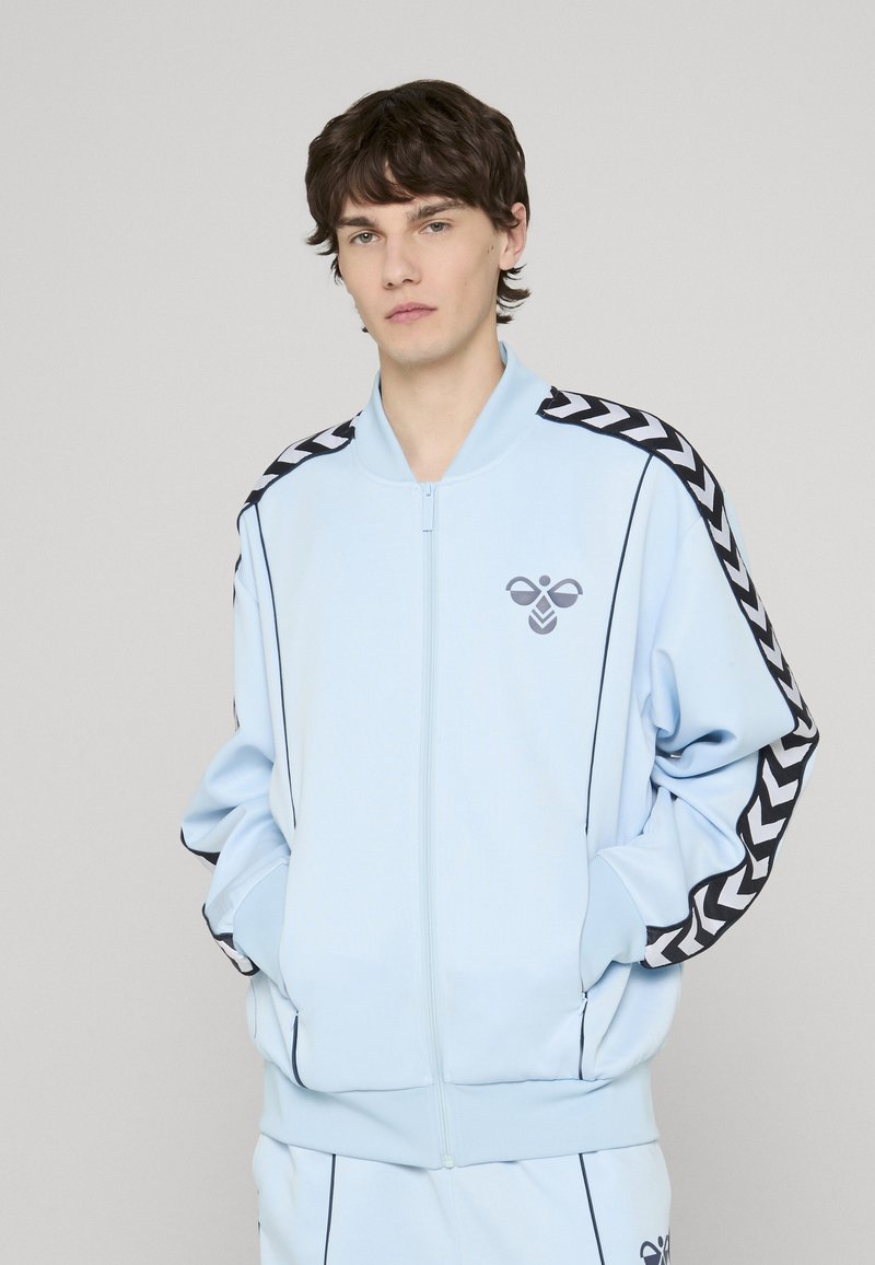 Veste bleu clair à zip fabriquée en tissu doux ; comporte des rayures en zigzag noires sur les épaules et un petit logo graphique sur la poitrine.