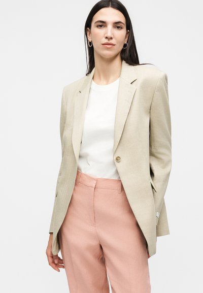 Γυναίκα με ίσια σκουρόχρωμα μαλλιά φοράει μπεζ σακάκι, λευκό μπλουζάκι, ροζ ψηλόμεσο παντελόνι και κρίκους σκουλαρίκια, στέκεται μπροστά σε απλό φόντο.