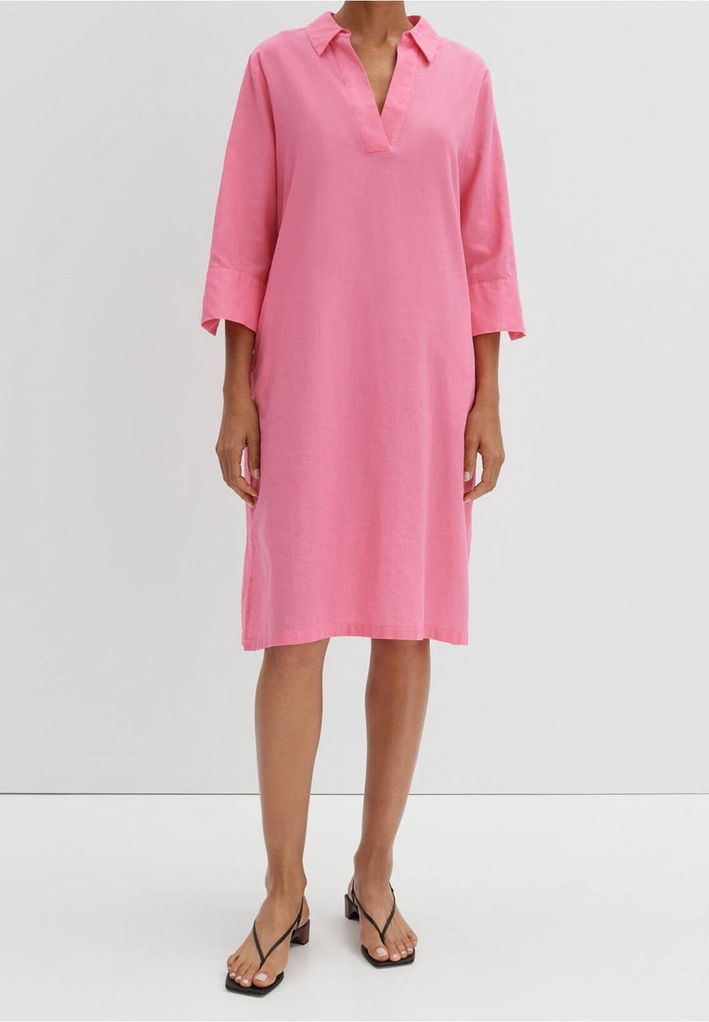 Kvinna i en knälång rosa klänning med V-ringning och trekvartsärmar, samt svarta öppna klackskor mot en vit bakgrund.