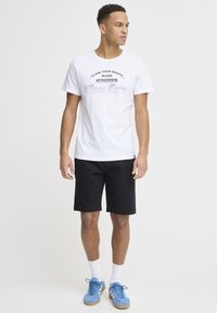 Hvit bomulls T-skjorte med trykket tekst, sammen med svarte shorts. Skoene er blå med en semsket tekstur. Modellen står i en nøytral posisjon.