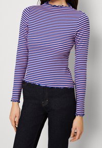 Długo rękawowy top w różowo-niebieskie paski, prążkowana tekstura, falbaniasty dół i mankiety, dopasowana sylwetka, noszony z ciemnoniebieskimi dżinsami.