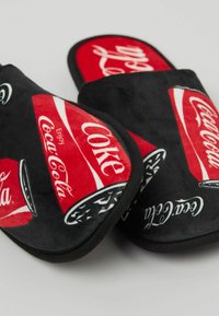 Červené a černé pantofle s designem hrnků Coca-Cola, vyrobené z měkké látky s texturovaným povrchem a černým lemováním podél okrajů.