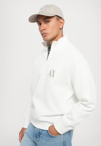 Vit zip-up sweatshirt med hög krage, med en grå logotyp på vänster bröst. Parat med ljusblå jeans och en beige keps.