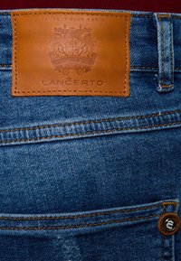 Džíny z denimu s hnědým koženým štítkem s vyraženými detaily a logem "LANCERTO", zdůrazněné oranžovým prošíváním a kovovým knoflíkem.