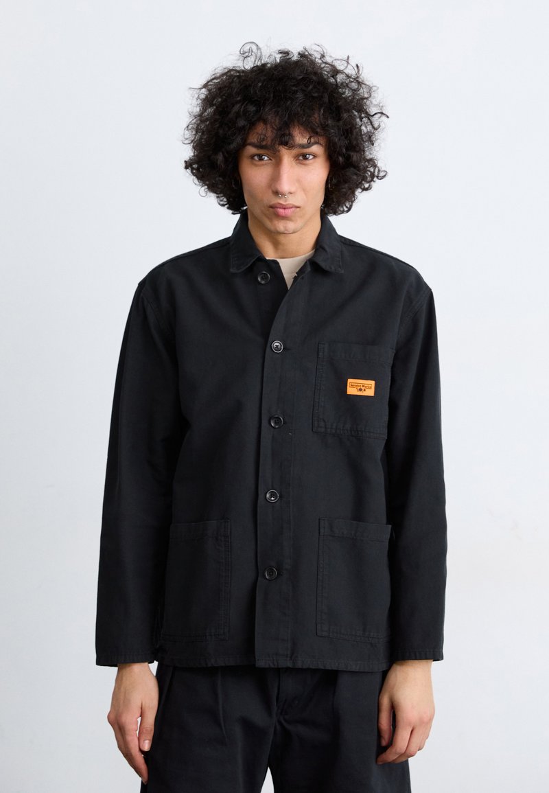 Black cotton work jacket featuring a button-down front, two front pockets, and an orange label on the left chest. Classic collar design.