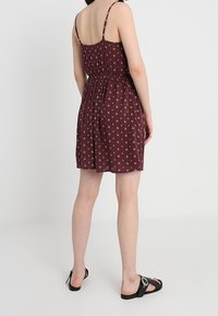 Robe d'été bordeaux avec une taille cintrée, des bretelles spaghetti réglables et un petit motif floral blanc. Portée avec des sandales à semelle noire.