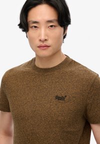 Camiseta de manga corta marrón hecha de tela texturada, con un escote redondo y un pequeño logo negro en el lado izquierdo del pecho.