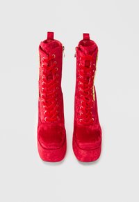 Bottes ankle en velours rouge avec un design plateforme, des finitions en ton or, des lacets à l'avant et une fermeture éclair sur le côté pour un enfilage facile. Texture douce et forme distincte.