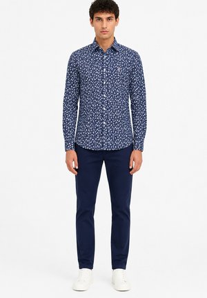 Jeune homme debout portant une chemise bleue à imprimé floral à manches longues, un pantalon bleu foncé et des baskets blanches sur un fond blanc uni.