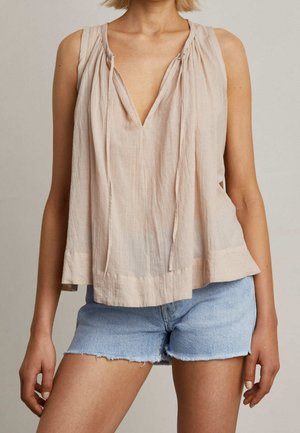 Femme portant un chemisier beige sans manches avec un col en V profond et un short en jean bleu clair aux bords effilochés.