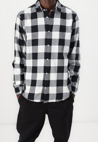 Camicia a maniche lunghe a scacchi bianco e nero con bottoni, realizzata in un tessuto testurizzato, indossata con pantaloni neri.