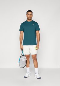 Teal idretts t-skjorte, hvite shorts, holder en tennisracket og en gul ball. Hvite sko med blå detaljer. Enkle, funksjonelle sportsklær.