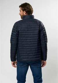 Marineblå puffjakke med horisontal quilting, høy krage og glatt tekstur, kombinert med blå jeans. Detaljert søm synlig.