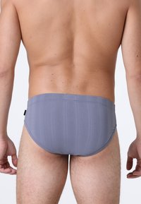 Vista posteriore di un uomo che indossa un costume da bagno grigio con sottili righe verticali, in piedi contro uno sfondo chiaro e neutro.