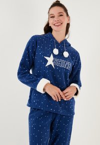 Navy plave pidžame s kapuljačom i uzorkom zvijezda, sa bijelim natpisom "SHINE", pahuljastim manžetama i pom-pom detaljima. Mekani, teksturirani materijal.