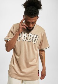 Tricou sportiv din plasă bej cu logo-ul "FUBU" în litere crem și negre. Prezintă mâneci scurte cu manșete în dungi și etichetă pe lateral.