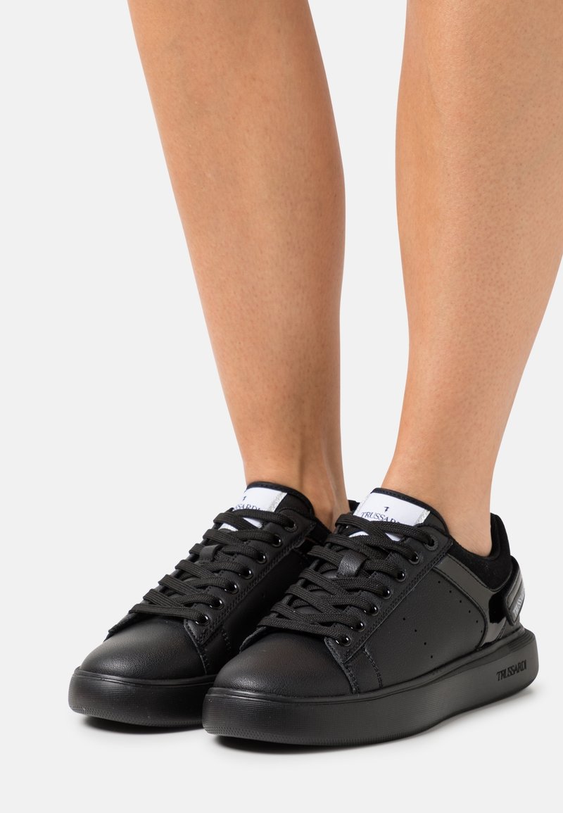 Personne portant des baskets basses noires avec des lacets noirs et des accents blancs sur la languette, se tenant sur une surface blanche.