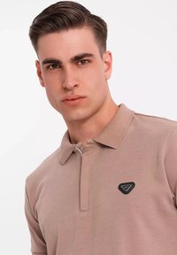 Polo de color marrón claro con cuello de media cremallera, mangas cortas y un parche de logo negro triangular en el lado izquierdo del pecho. Textura suave.