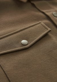 Tejido marrón texturizado con un bolsillo en el pecho y un botón a presión plateado. El material tiene un diseño acanalado, que muestra una construcción robusta.