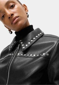 Zwarte leren jas met een hoge kraag, zilveren studs langs de voorkant en kraag, en een ritssluiting aan de voorkant.