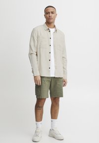 Camisa beige de botones con dos bolsillos en el pecho, combinada con pantalones cortos de cargo en color verde oliva. Camiseta blanca debajo, calcetines blancos y zapatillas beige.