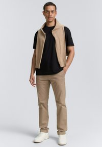 Beige sweatervest over en sort t-shirt, parret med lysebeige bukser og hvide sneakers. Outfittet har et rent, afslappet look.