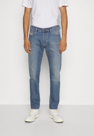 Man die medium-wash blauwe spijkerbroek, wit T-shirt en witte sneakers draagt, staande tegen een eenvoudige witte achtergrond.