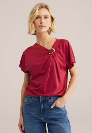 Blondínka v červenom topu so zlatou prackou na ramene a modrých džínsach, stojaca na neutrálom pozadí.