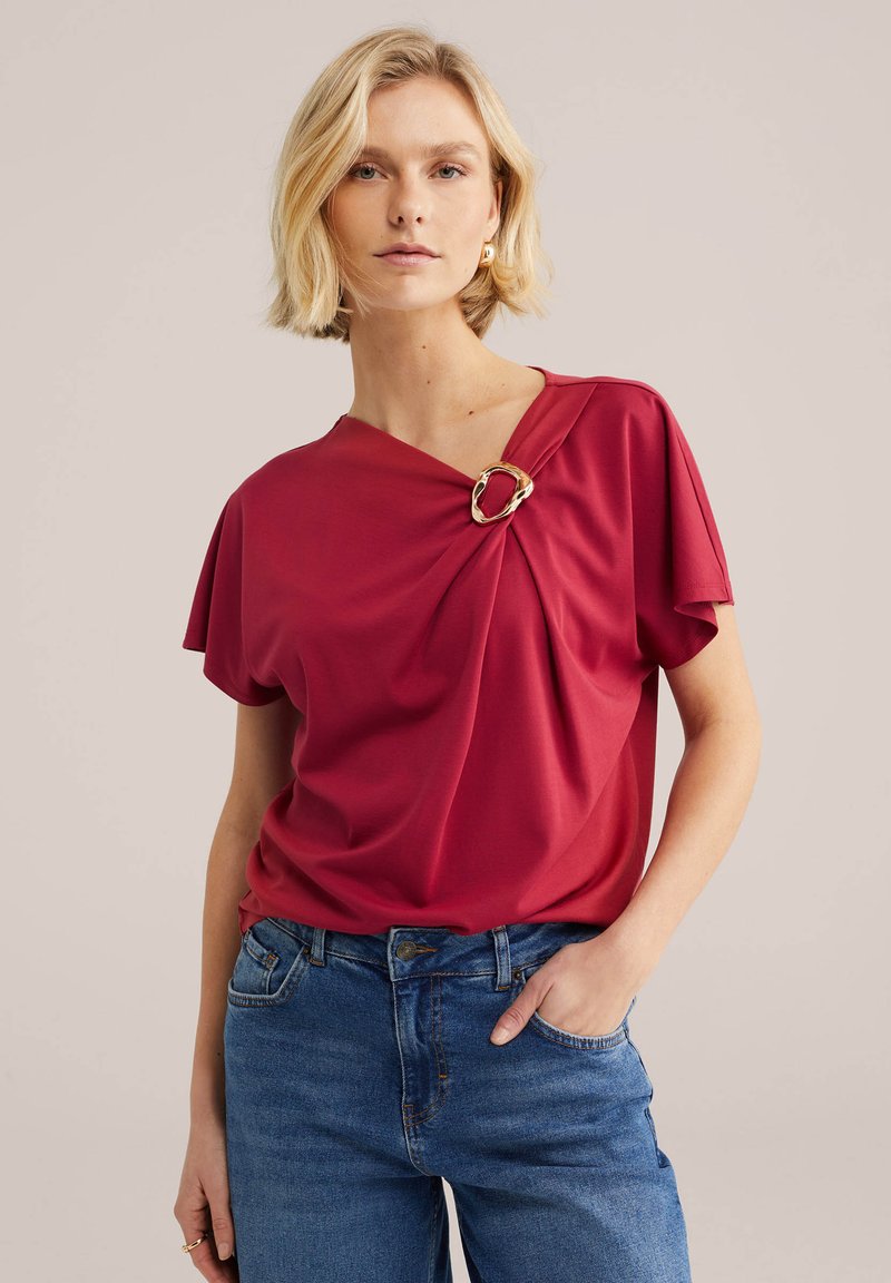 Femme blonde portant un haut rouge avec un détail boucle dorée sur l'épaule et un jean bleu, debout devant un fond neutre.