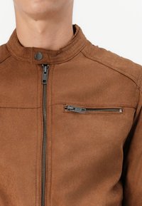 Giacca in finta pelle marrone con colletto a imbuto, chiusura frontale con zip e tasca sul petto con dettagli in zip argentata. Tessuto morbido.