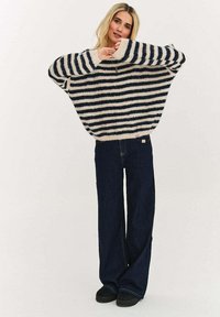 Przewymiarowany dziany sweter z granatowo-kremowymi poziomymi paskami, z szerokim dekoltem i opuszczonymi ramionami, zestawiony z ciemnymi rozszerzanymi jeansami.