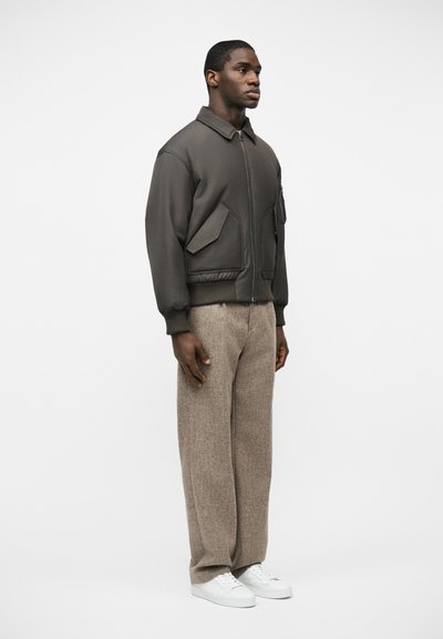 Homme debout, portant une veste zippée gris foncé avec de grandes poches, un pantalon à jambes larges beige et des baskets blanches, sur un fond blanc uni.