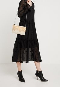 Czarna sukienka midi w groszki z falbanką u dołu, zestawiona z czarnymi teksturowanymi botkami za kostkę. W ręku trzymana jest beżowa torebka zdobiona koralikami z łańcuszkowym paskiem.