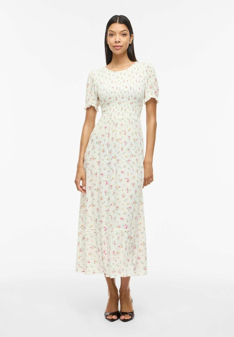 Femme debout dans une robe midi blanche à motifs floraux avec manches bouffantes et ourlet à volants, portant des talons ouverts noirs sur un fond uni.