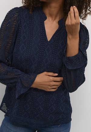 Femme portant un chemisier en dentelle bleu marine avec des motifs en losange et des manches bouffantes, vue du cou aux hanches, une main près du visage.