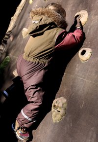 Gyermek egy texturált mászófalon mászik, barna-zöld párnázott dzsekit visel szőrmebéléssel ellátott kapucnival és barna mászó nadrágot.