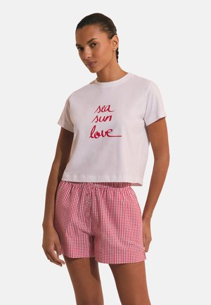 Kobieta ubrana w biały krótki t-shirt z czerwonym napisem "sea sun love" i czerwono-białe kraciaste szorty, stojąca na jednolitym tle.