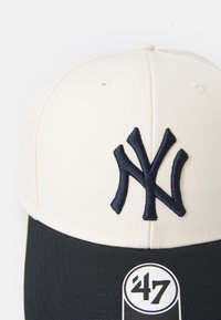 Gräddfärgad och marinblå basebollkeps med broderad "NY"-logotyp, strukturerad design, böjd skärm och en märkesetikett med '47-logotyp.