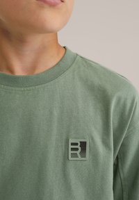 Svetlo zelena bombažna majica s krožnim ovratnikom, z kvadratnim črnim logotipom na levem prsnem delu. Mehak, teksturiran material.