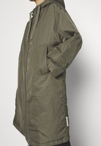 Parka verde oliva con cappuccio, con zip frontale completa, due tasche laterali e polsini con bottoni a pressione. Realizzata in un materiale liscio e leggero.