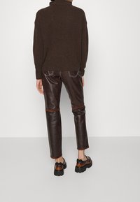 Personne portant un pantalon en cuir marron foncé avec des coutures contrastantes, un pull marron, et des chaussures grosses bicolores marron et noir, debout devant un fond uni.