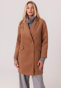Dwurzędowy płaszcz w kolorze camel, wykonany z mieszanki wełny, z kołnierzem typu notch, przednimi kieszeniami i luźnym krojem, noszony na szarym golfie.