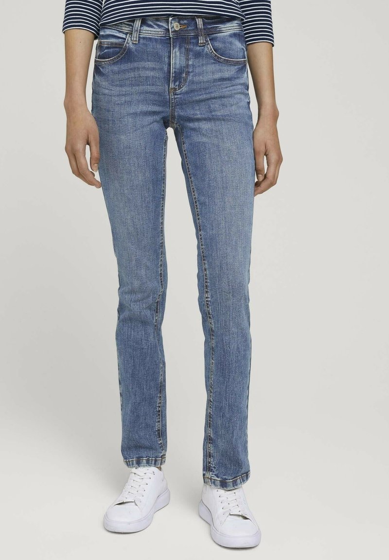 Personne portant un jean taille mi-haute, bleu clair, coupe droite, avec des baskets blanches et une chemise rayée à manches longues, debout devant un fond uni.