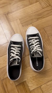 Schwarze Canvas-Sneaker mit niedrigem Schaft, weißer Gummikappe, weißen Schnürsenkeln und schwarzem Innenfutter, auf einem Holzboden platziert.