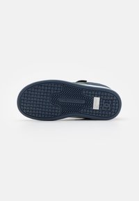Kék sportcipő talp, textúrált tapadási mintával, DC Shoes logóval és egy téglalap alakú fehér címkével a sarok részen, fehér háttér előtt.
