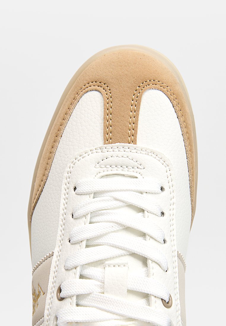Zapatilla blanca con base de cuero suave, detalles en ante marrón, punta redonda y cordones blancos planos. Logotipo sutil en el costado.