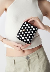 Portafoglio nero con pois bianchi, dotato di una cerniera e accessori in oro. Materiale testurizzato per una maggiore durata. Design rettangolare compatto.