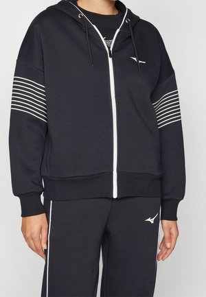 Persona che indossa una felpa nera con zip e strisce bianche sulle maniche e pantaloni neri abbinati con strisce bianche e logo.